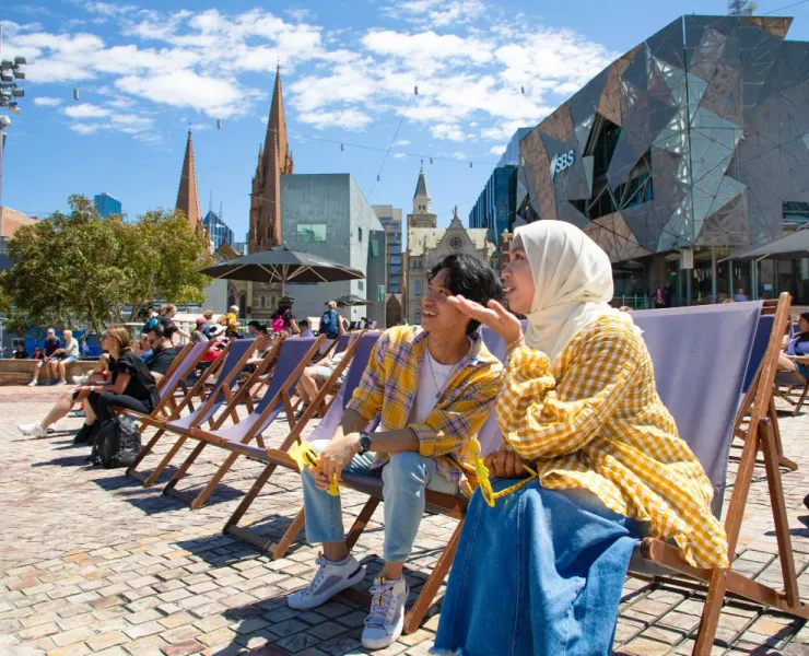 Two young people sit on chairs in Federation Square in Melbourne. It appears as though they are watching something either on screen on on the stage ahead.